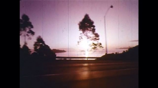 UNITED STATES: 1950s: sunset by road. Man and lady enjoy picnic in countryside. View of car and coast. People and car at sunset