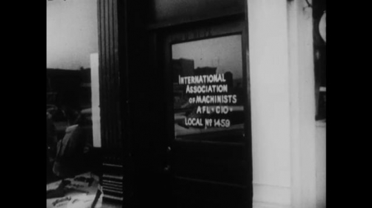 UNITED STATES: 1960s: Shop door sign: International Association of Machinists. Man enters shop. Employees leave through employees entrance