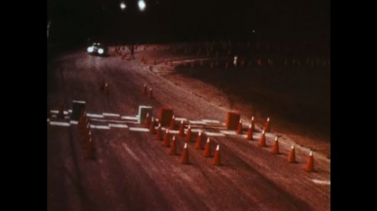 UNITED STATES: 1970s: car drives on track at night. Car swerves. Car hits cones. 