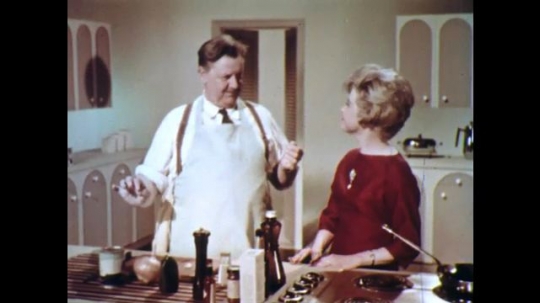 UNITED STATES: 1960s: lady talks to man in studio kitchen. Lady passes bowl to man