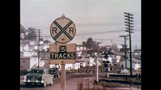 UNITED STATES: 1970s: railway crossing sign posts. Road above railway lines. Train on tracks