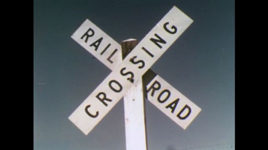 UNITED STATES: 1970s: railway crossing sign. Car approaches junction. Railway tracks. Bus crosses tracks
