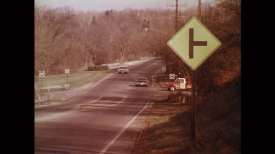 UNITED STATES: 1980s: cars drive along road. Sign on side of road. Vehicle pulls onto road. Close up of sign. View through windshield
