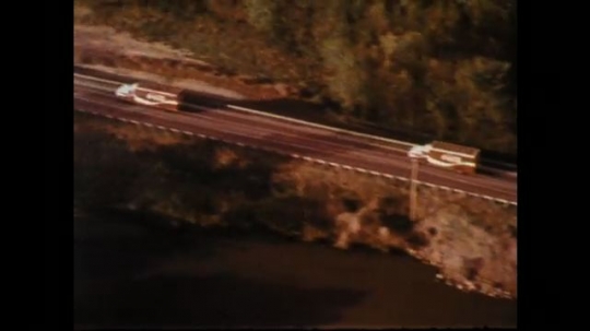 1970s: Aerial of trucks. Truck on bridge. Driving on bridge. Cabin. Truck on road. Man driving. Man exits store. Man on bench. Man driving. Hands on guitars. Man singing. Man dusting. Truck driving. 