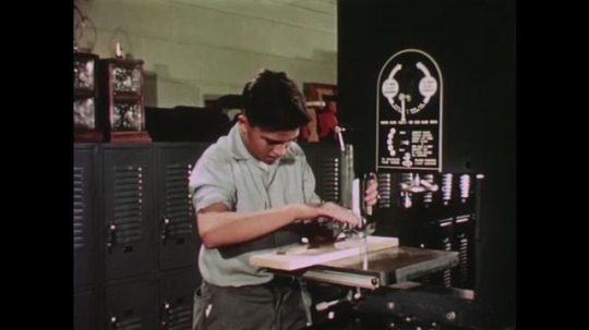 UNITED STATES: 1950s: boy turns handle to adjust jigsaw machine. Boy checks note book. Boy cuts wood
