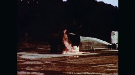 1970s: Firefighters spray hoses on flames at one end of a horizontal cylindrical storage tank. 