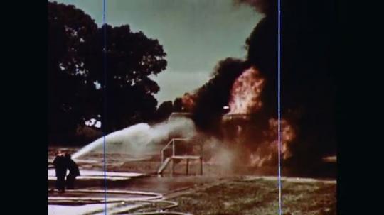 1970s: A vertical tank and a horizontal tank adjacent to each other outdoors are in flames with great clouds of black smoke. Firefighters train their hoses and hold their ground on the fire.