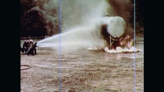 1970s: A horizontal cylindrical tank on a scaffoldlike mount is in flames. Firefighters train hoses on tank supports. The scaffold collapses from the heat and fuel in tank feeds the fire.