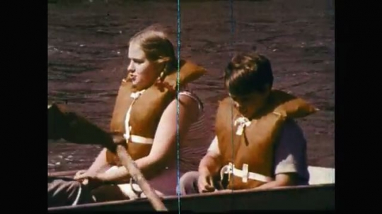 1960s: UNITED STATES: children on rowing boat. Man sits on cushion on boat. Man swims. Girl swims. Buoyancy aid on side of water