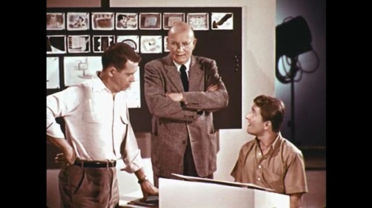 1960s: men stand around desk and talk near storyboard. man rests on moviola on soundstudio with lamp nearby.