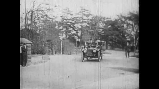 1910s: Car full of passengers driving. Car pulls up to building, men exit car. 