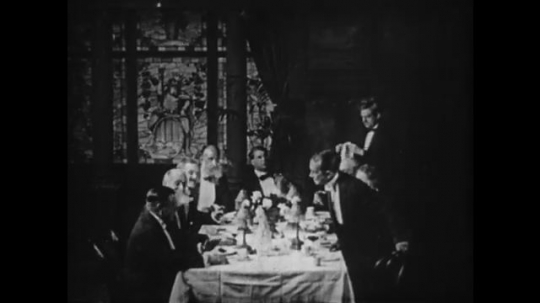 1910s: Man around dinner table, man stands. Intertitle. Men stand and toast. 