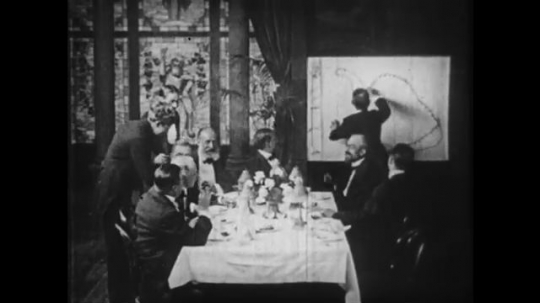 1910s: Men around dinner table, man drawing in background. 