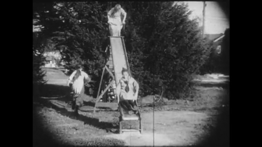 1910s: Group of girls play on a slide, another girl approaches a bench where some dolls have been left behind. She contemplates picking one up and eventually does.