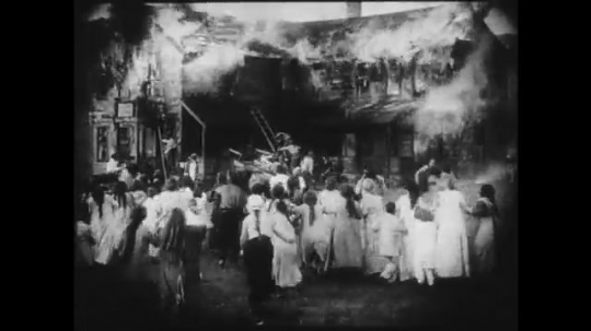 1910s: Large crowd panics outside of a burning building. A smaller group gazes at the destruction, some of them talk to a girl and a young man stares at her. Firefighters try to put out the flames.