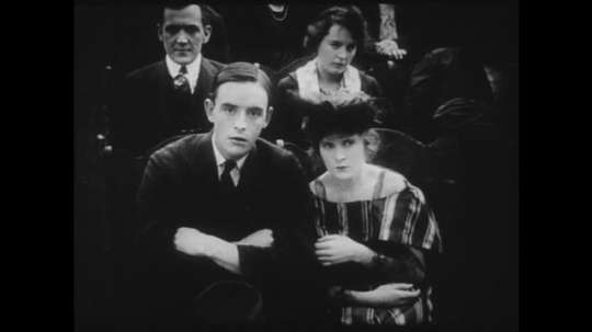 1910s: Young couple watches a movie in a theater, they watch the screen with apprehension. Movie shows man dying after being shot and woman fetching water from well.