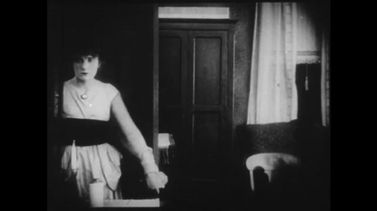 1910s: Young woman enters a parlor and overhears people outside concerned that they heard a gunshot, she looks guilty/worried. Woman walks around in living room.