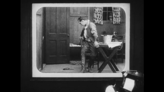 1910s: Movie screen shows man falling out of chair and crawling away, cuts to woman looking worried then walking off screen.