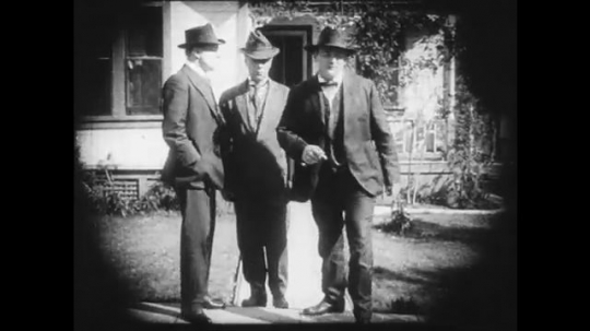 1910s: Group of three men on a sidewalk, two leave then the last one leaves and meets a different larger group of men. Young woman looks worried in a room.