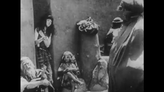 1910s: UNITED STATES: child knits in street. People in street. Lady looks through window. Women work