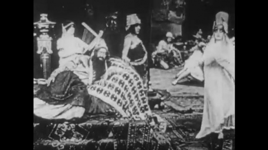 1910s: UNITED STATES: lady dances in street for man. Man plays music for leader on throne. 