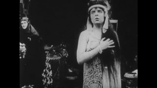 1910s: UNITED STATES: lady holds hand across her chest. Lady wraps cloak around girl. Man on throne
