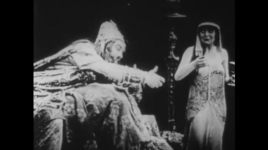 1910s: UNITED STATES: king sits with lady on throne. Lady encourages man to drink from goblet. Man pushes goblet away. Man drinks