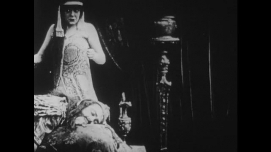 1910s: UNITED STATES: lady stands over body of man on throne. Lady raises sword above man