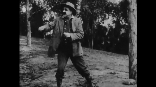 1910s: man with cowboy hat points, gets shot in the butt and throws rock at man with mustache and rifle. black cat jumps and runs away in woods.