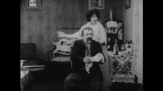 1910s: woman hands towel to man with mustache and leaves. woman argues and hits man with cowboy hat in the living room. titlecard.