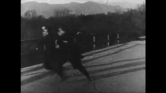 1910s: three men in police uniforms stand on bridge and fire guns as man with mustache drives motorcycle, shoots revolver and snags unconscious woman.