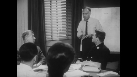 1950s: Engineer stands and talks to people in office meeting. Experts listen. Engineer puts on jacket and sits down.