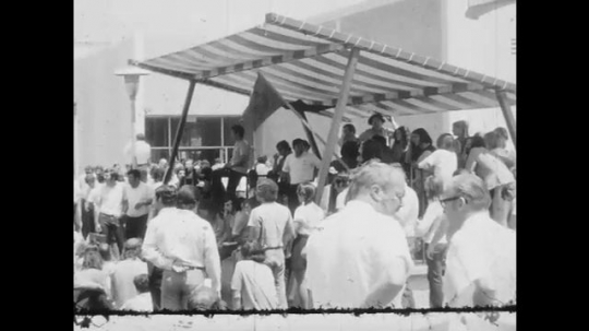 1970s: UNITED STATES: crowd gathers under awning. People gather outside building. People in park 
