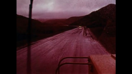 1970s: Vehicle moves slowly down slippery road, passes another truck. A man stands outside a pickup truck. Another vehicle approaches. 