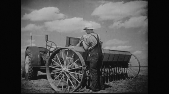1930s: Boy pours seeds into tractor. Close up, hands pouring seeds. Boy climbs onto tractor. Blades on tractor, tractor pulls away. 
