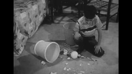 1950s: Boy on floor, cleans up broken lamp. Man in doorway, bends down. Man helps boy clean up lamp. 