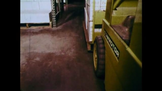 1970s: UNITED STATES: view of truck wheels on warehouse floor. Truck moves packages across warehouse. Man drives truck