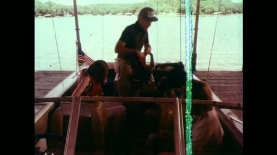 1970s: Man and three teens stand on a boat and and put on life jackets. Fire extinguisher. Coil of blue throw line. First Aid kit. Wooden paddle.  Yellow rope coil. Small anchor in bottom of boat. 