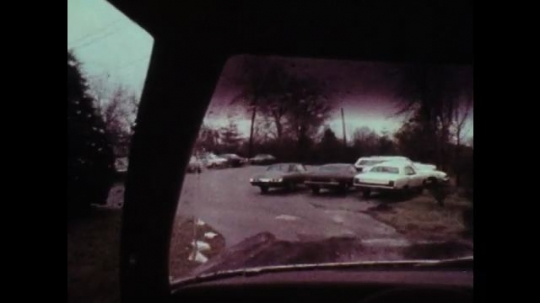 1970s: UNITED STATES: view through window of car. Wipers on window in rain. Cars in car park. Man winds down window. Man walks under umbrella