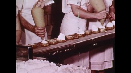 1960s: Bakery workers pipe whipped cream onto pastries, man and boy watch. Boy smiles. Bakery worker breaks up large chocolate bar, puts pieces into bowl. Melted chocolate pours into bowl.