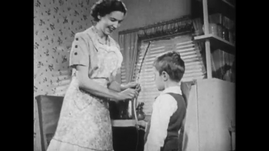 1950s: Mother helps boy fix loose bow. Boy looks at bow. 
