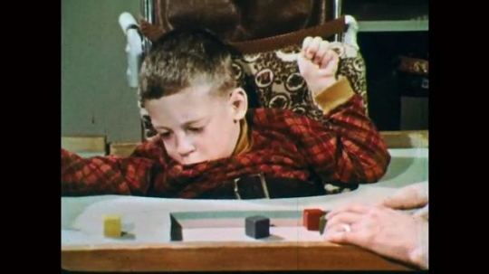 1970s: Boy in wheelchair works with blocks. Hand points to blocks. Hand on table. Model of human brain with lights flashing.