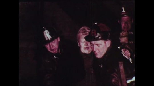 1970s: Firefighters help people down fire escape. Man in uniform shows newspaper to another man with headline concerning retirement home fire.