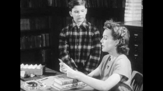 1940s: Woman sits at desk, boy stands by desk, woman hands card to boy, talks to boy. Boy takes card, talks, smiles, walks away. Woman moves books to side of desk. Boy exits library.