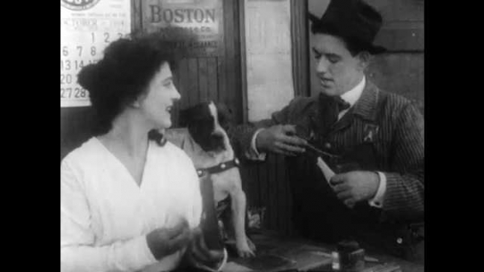 1910s: Man in hat talks to woman while woman pets dog. Man in overalls picks up oil can. Another man walks out of station, smiling.
