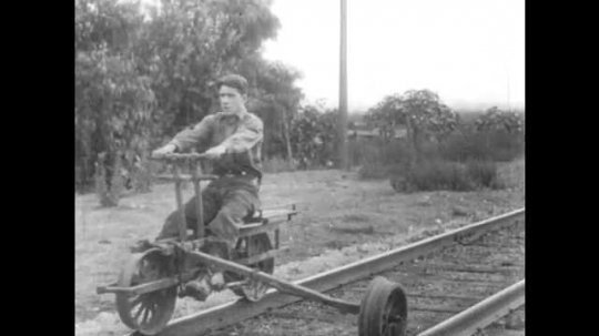 1910s: Train on track passes through rural area. Wounded man on ground, boy moves pedal cart to man. Boy lifts man