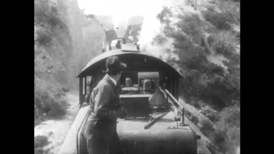 1910s: Man on train looks back, then runs to front. Boy on push cart stops moving, heads into train station. He hands another boy piece of paper, then points at it. Woman reads paper. Boys talk.