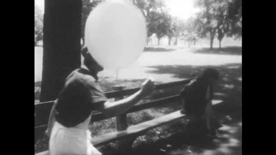 1960s: woman in dress and black hair sits on bench as man in apron and cap holds a balloon, walks back and forth on path and talks loudly in city park with trees.