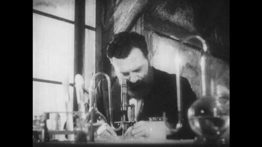 1930s: 1860s reenactment. A bearded man looks through a microscope, then holds beakers up to the light. Wine bottles sit next to test tubes on a table.