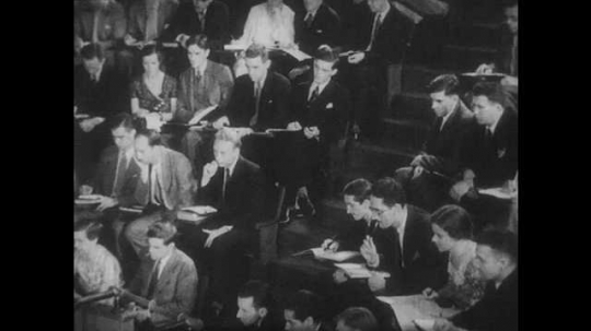 1930s: Students sit in a lecture hall. A man lectures at the front of the room.